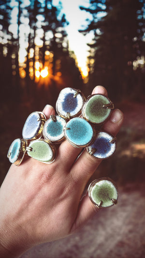 Photo of a hand in front of a sunset forest with multiple large and colorful rings on each finger