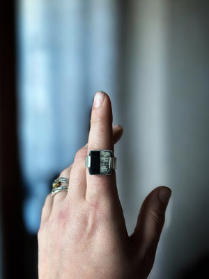 hand in front of a blurred background wearing a white and black silver ring.